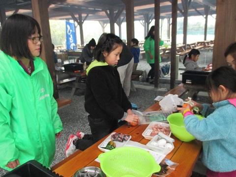肉や野菜などの食材やボウルなどの調理器具が用意された調理場で、関係者に見守られながら調理をしている子どもたちの写真