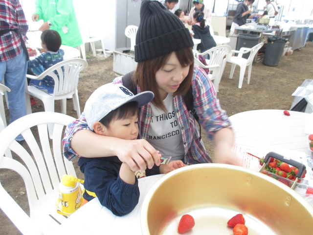 ニット帽を被った女性とキャップの帽子を被った子どもが、イチゴを大きな鍋に一緒に投入している写真
