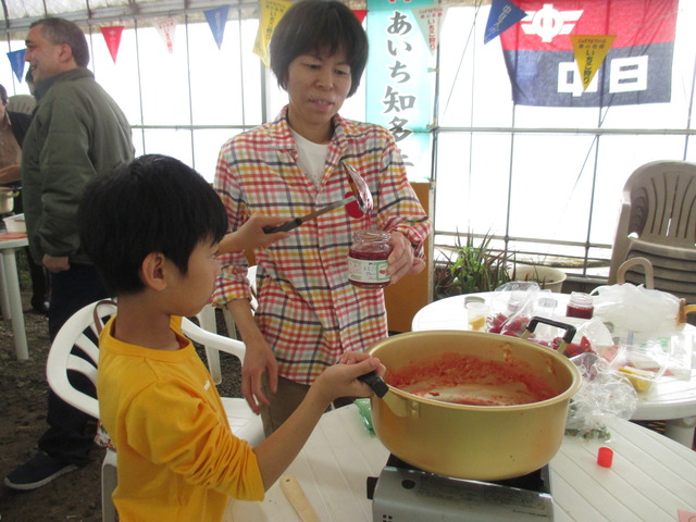 完成したイチゴジャムをお玉を使って鍋から瓶へ運んでいる男の子と、瓶を手に持って支えている女性の写真
