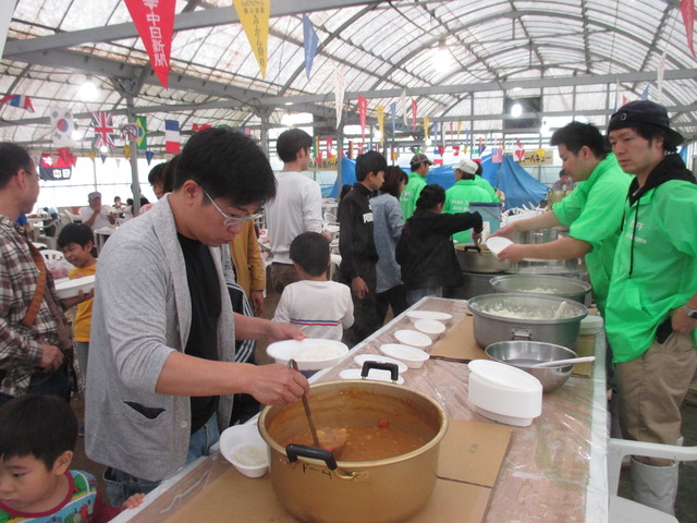 大きな鍋に白飯やカレーが用意されたビニールハウスの会場が、器にカレーをよそっている人や列を作って並んでいる人たちで賑わっている写真