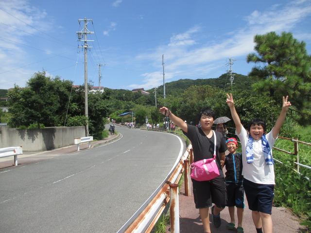 左側に2車線の道路がある横の歩道を、手をあげたり、ピースサインをしながら歩いている3人の男の子たちの写真