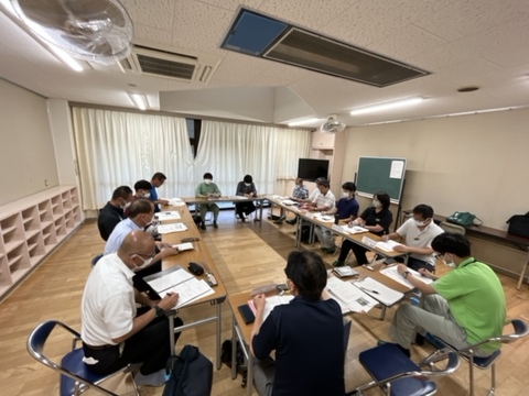 左側に棚、右側に黒板などが設置された室内で、長机をロの字型に設置し、関係者たちが席に座り会議を行っている様子の写真
