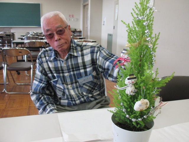 貝殻などを使って装飾を施した円錐状の樹形の鉢植えと、椅子に座って鉢植えの方を見つめている年配の男性の写真