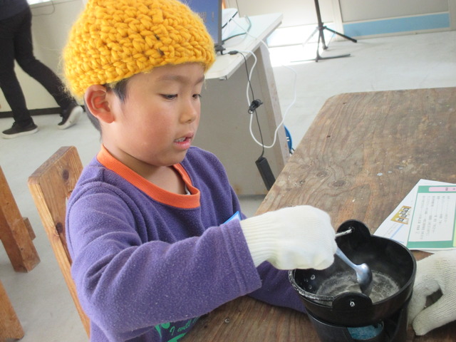 白く浮かび上がった塩を見つめながら小さな鍋をスプーンでかき混ぜている男の子の写真