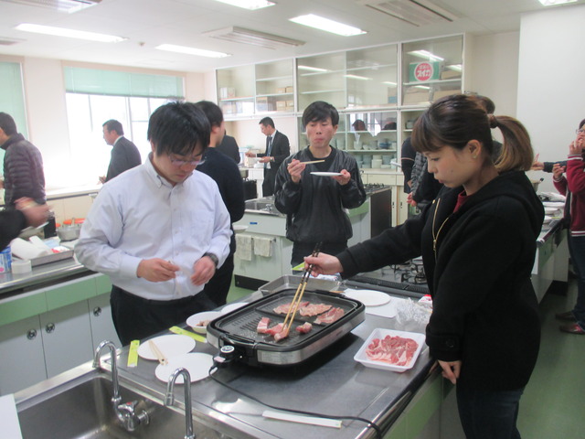 菜箸を使いプレートでお肉を焼いている女性、その周りで男性が試食をしている写真