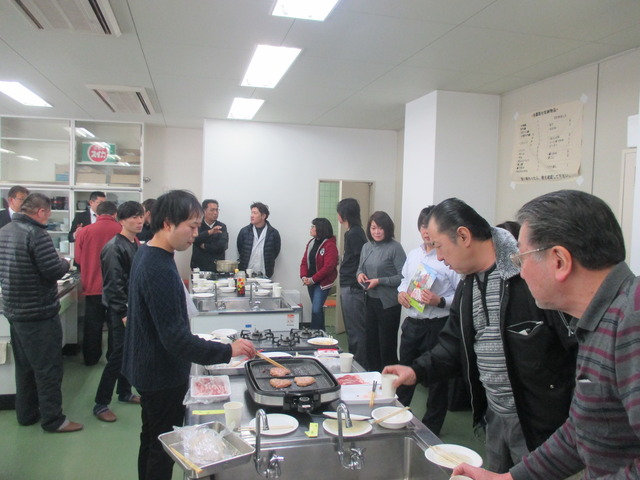 男性1人がプレートでお肉を焼き、調理室に集まった男女の参加者達が試食をしている様子の写真