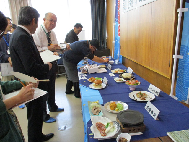 飲食店や旅館名が書かれた名札の前にいろいろな種類の料理が並べら、それを審査している関係者達を写した写真