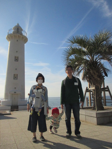 白色の野間灯台と海をバックにして、赤い帽子を被った子どもを中心に手を繋いで記念撮影をしている家族の写真
