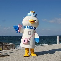 青雲の下、広い海をバックにアスファルトに立っているマスコットキャラクター「のまっキー」の写真