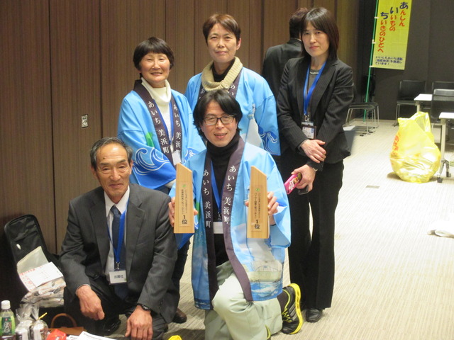 前列にスーツ姿の男性と「いいともあいち交流会2017 バイヤー試食人気コンテスト1位」と書かれた木製のトロフィーの様な物を両手に持っている男性が膝をつけて座り、後方に法被を着た2名の女性とスーツ姿の女性が立ち、記念撮影をしている写真