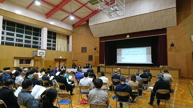 ステージに資料が映し出されたスクリーンが設置され、ステージ下の左側の関係者が手元の資料を見ながら話をし、パイプ椅子に座っている多くの参加者が話を聞いている様子を体育館の後方から写した写真
