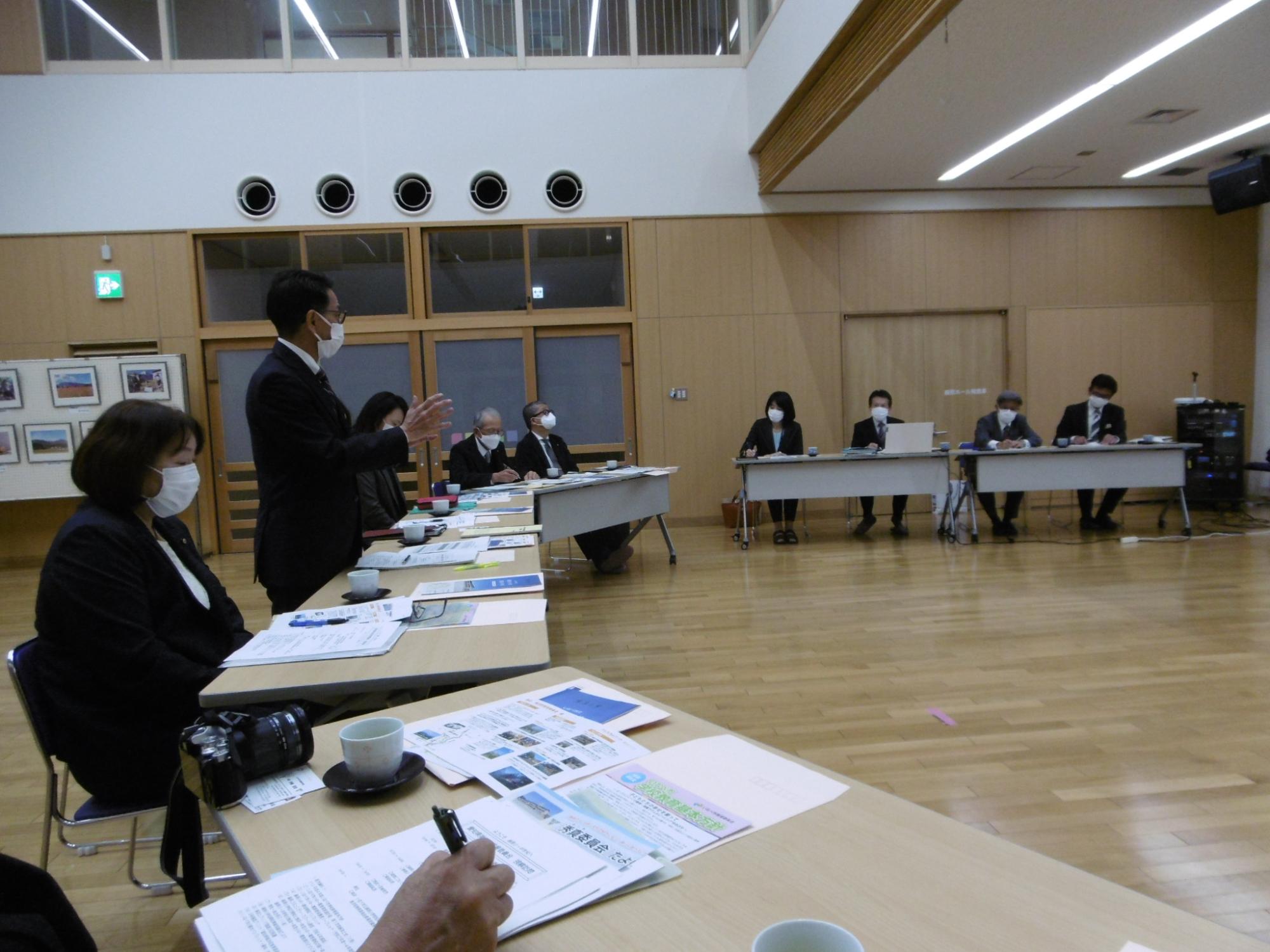 大きなホールのような場所に扇状に長机が設置され各2名ずつ席につき、中央付近の席の男性が立って話をしている様子の写真