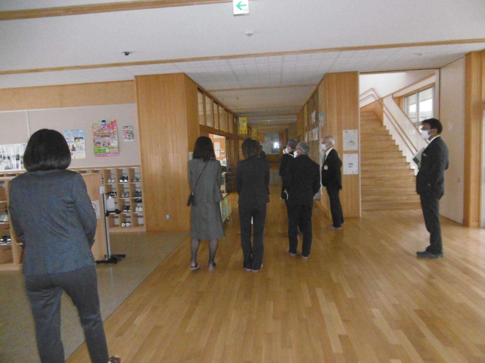 床や壁が木目調になっている校内で、靴箱付近を視察しているスーツ姿の関係者10名弱の写真