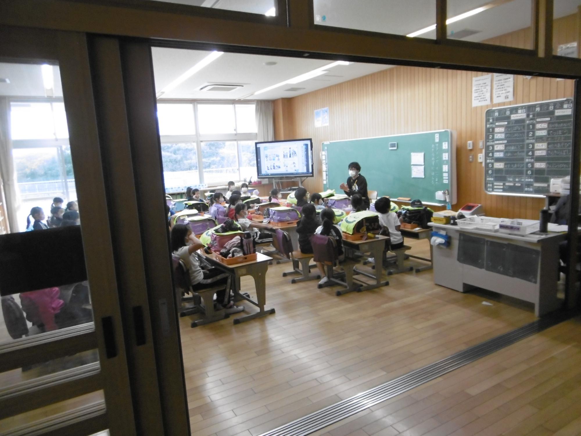 教室の廊下側が壁の代わりにフルオープンできるガラス張りの扉になっており、全体が見渡せる教室内で生徒が席につき教卓の前に立つ先生が話をしている様子の写真