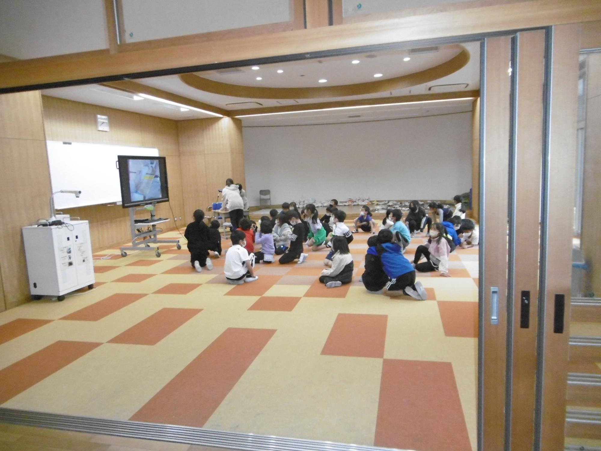 床にクリーム色とオレンジのタイルが貼られたホールのような場所で生徒たちが床に座り、正面のモニターに写された先生の手元を写すカメラの映像を見ている授業風景の写真