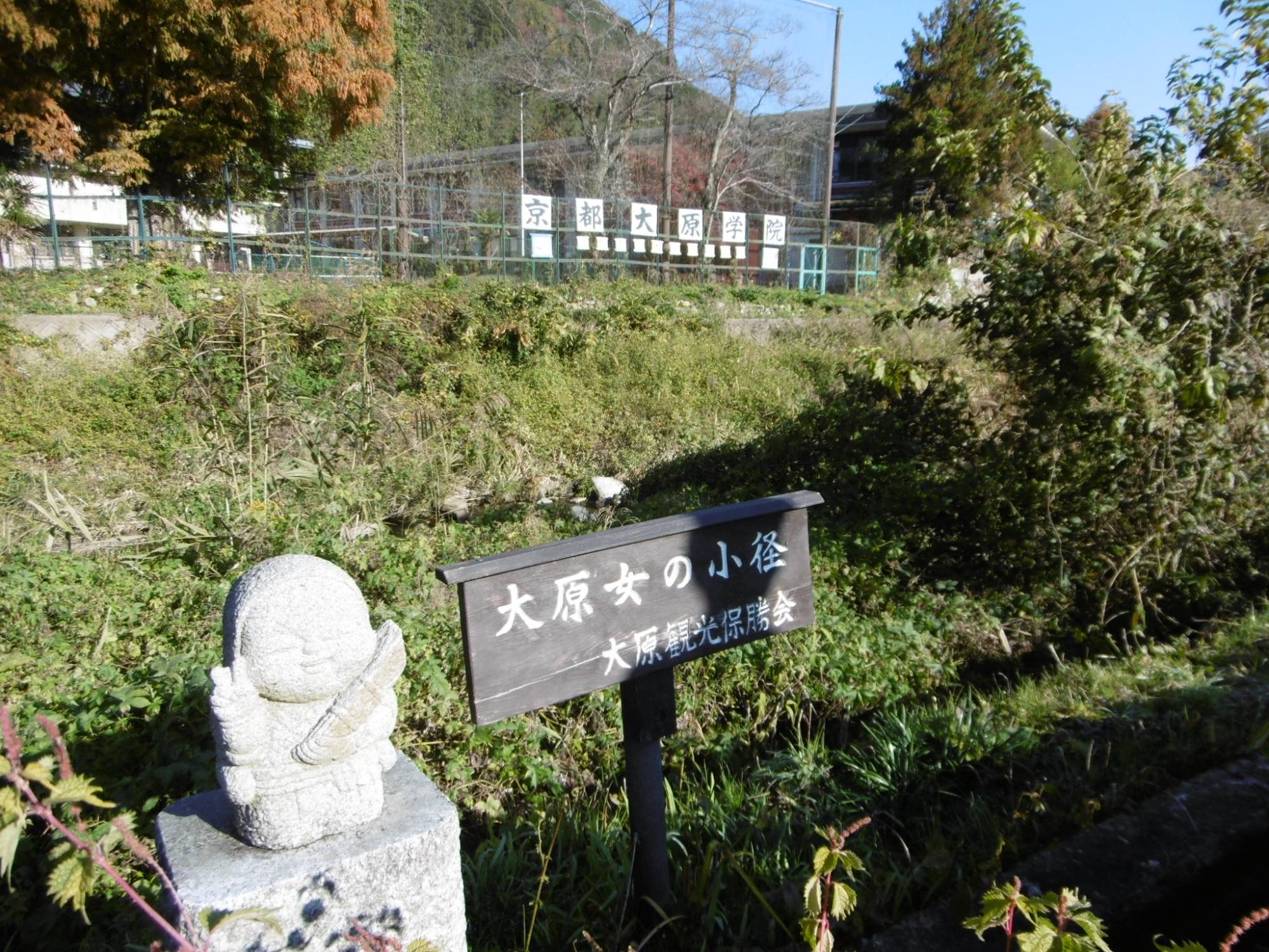 大原女の小径と書かれた看板とピースをしたお地蔵さんが並んで立ち、その奥に土手と小川をはさんで京都大原学院と書かれた看板がフェンスに設置されている写真