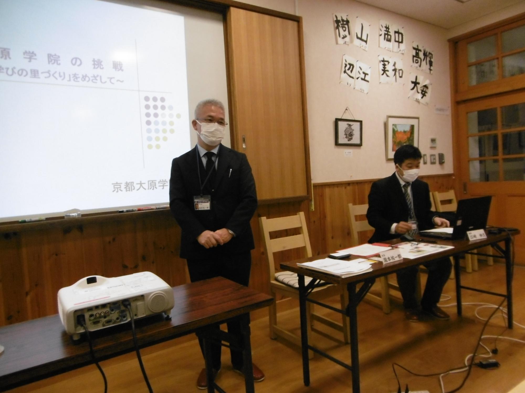 資料が写されたプロジェクターの横に立つ男性と、その横でパソコンを操作する男性の写真