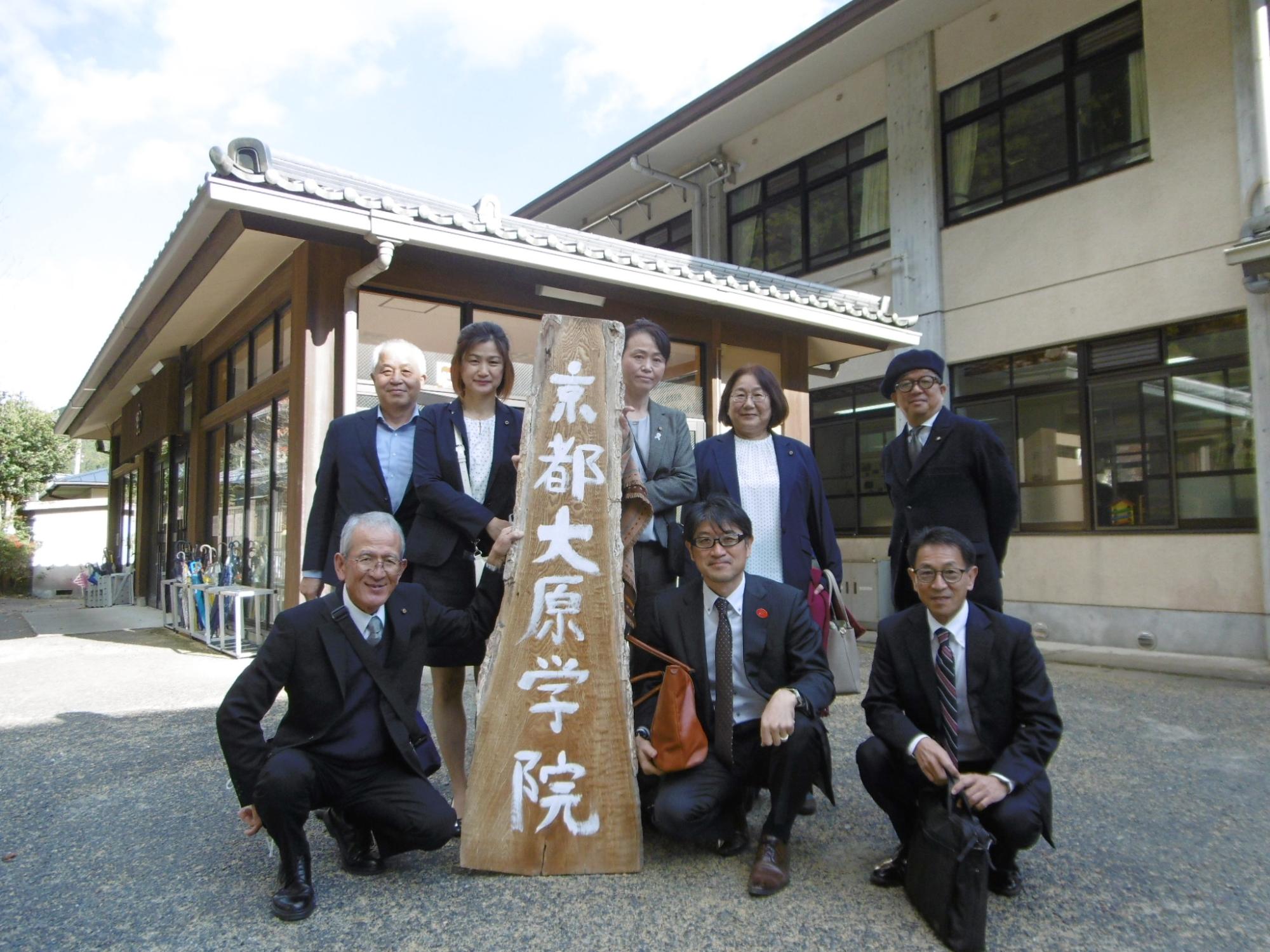校舎の前で「京都大原学院」と書かれた木製の看板を持って記念撮影する男性5名女性3名の関係者の方々の写真