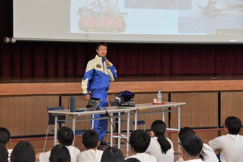 体育館の舞台前で、右手にマイクを持った磯部選手が長机が設置された席の横に立ち、体育座りをしている生徒たちに向かって話をしている様子を生徒たちの後方から写した写真