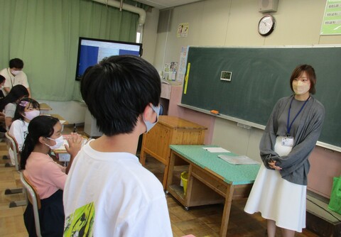 右側の最前列の男の子が立っている様子と向かい側に立って話を聞いているシェンさんの写真