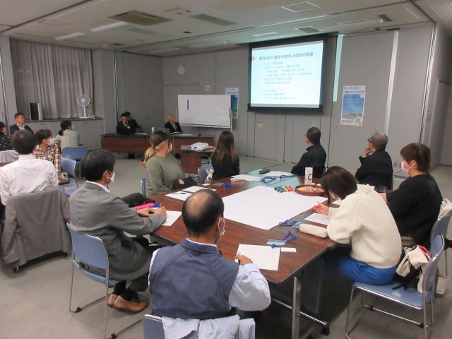 資料が置かれた大きなテーブルを囲むように椅子が配置された部屋で、着席した関係者の方々が、部屋の正面に設置されたプロジェクタースクリーンの方を見ている写真