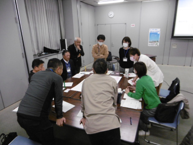 大きなテーブルを9名の関係者たちが囲むように集まっており、テーブル中央に置かれた大きな資料を見ながら話し合いを行っている様子の写真