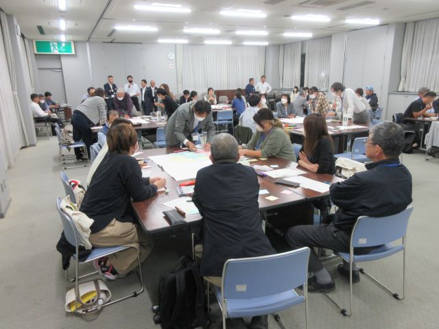 間隔をあけて大きなテーブルが複数用意された部屋で、グループごとに分かれて着席した関係者たちがテーブル上の大きな紙を使って話し合いを行っている様子の写真