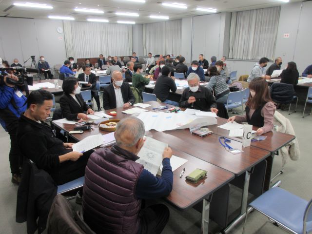 間隔をあけて大きなテーブルが複数用意された部屋で、グループごとに分かれて着席した関係者たちが手元の資料を見ながら話し合いを行っている様子の写真