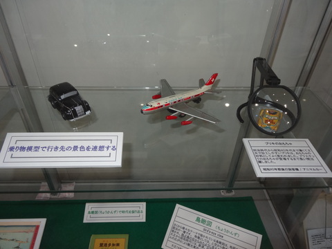 黒の車や飛行機の模型、虫眼鏡のレンズの先に黄色の車の様なものが映っているおもちゃが並べて展示されている写真