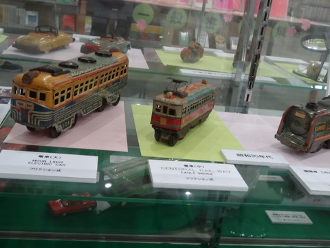 説明文とともに赤色や黄色、黒色など、カラフルな電車のおもちゃが複数展示されている写真