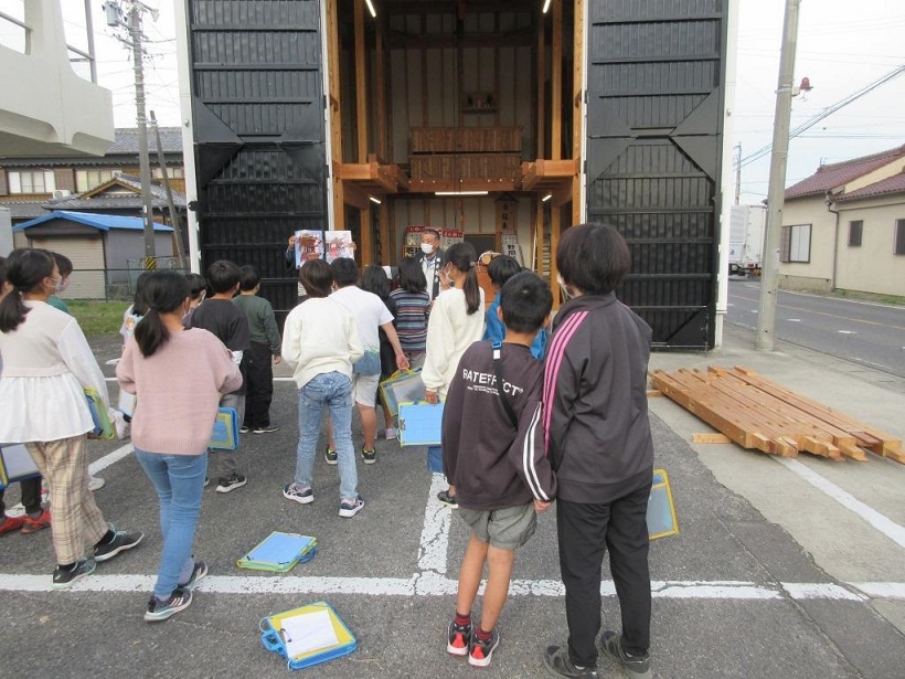 さまざまな道具が保管されている巨大な倉庫の前に子どもたちが集まり、倉庫入り口近くに立つ関係者の方を向いている写真