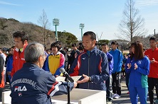 屋外で演壇を挟んで関係者の男性と参加者の男性が向かい合って立ち賞状を受け取っている表彰式の様子の写真