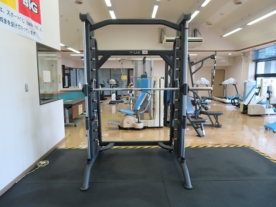 様々な器具が並んでいるトレーニング室の一角に、金属の棒が組み合わさっている形状をしたスミスマシンが1台設置されている写真