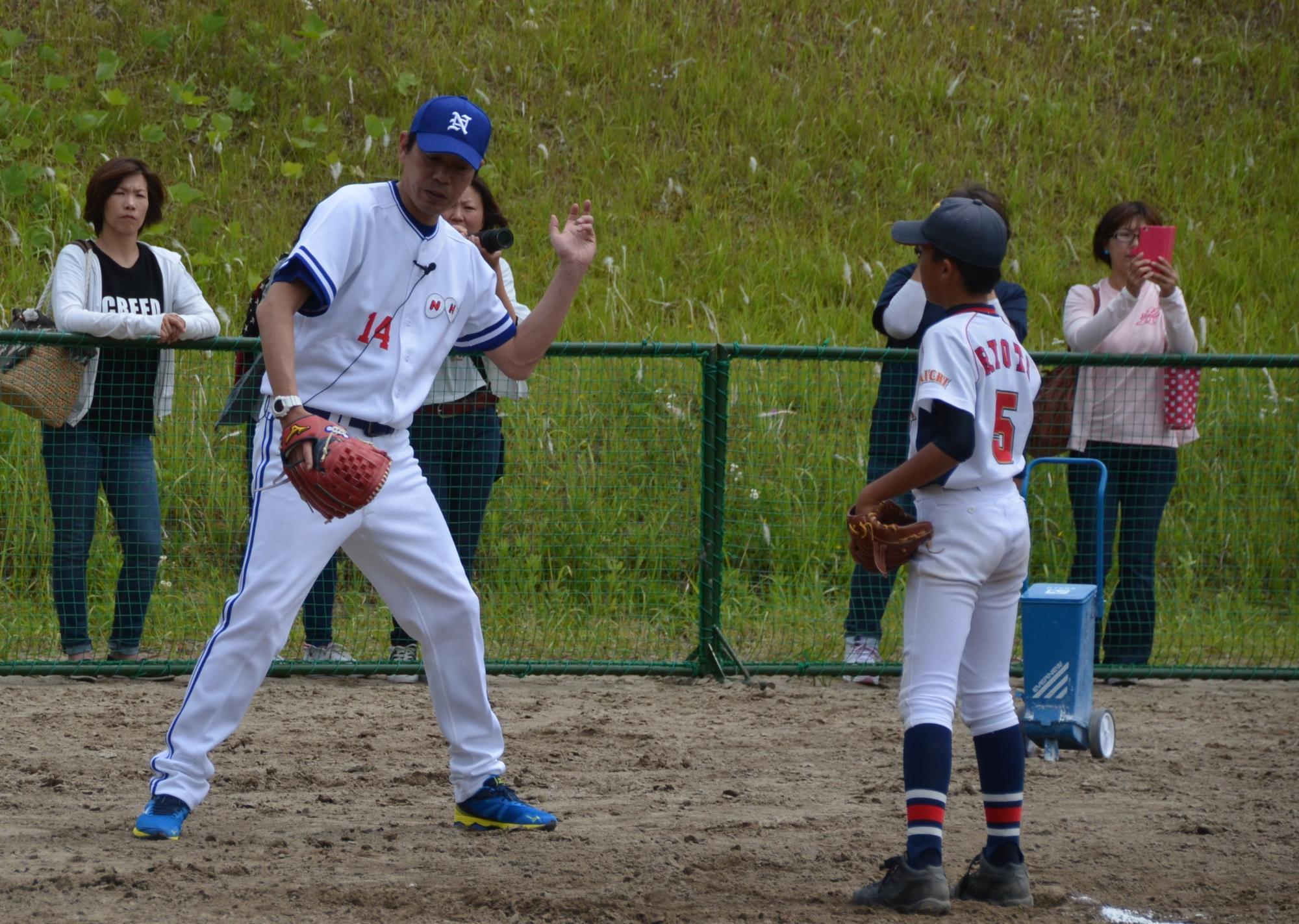 今西選手が背番号5をつけた選手に投げ方を指導している様子をフェンス越しに女性がみている写真