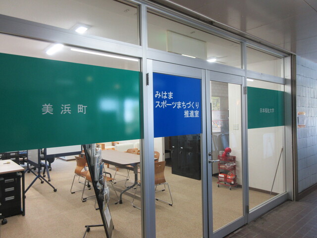 窓に「美浜町 みはまスポーツまちづくり推進室 日本福祉大学」と書かれ、室内にテーブルと椅子が設置された推進室室外から写した写真
