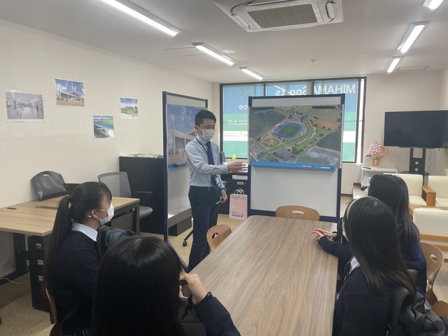 前で職員の方が説明するのを学生さん4人が椅子に座って話を聞いている写真