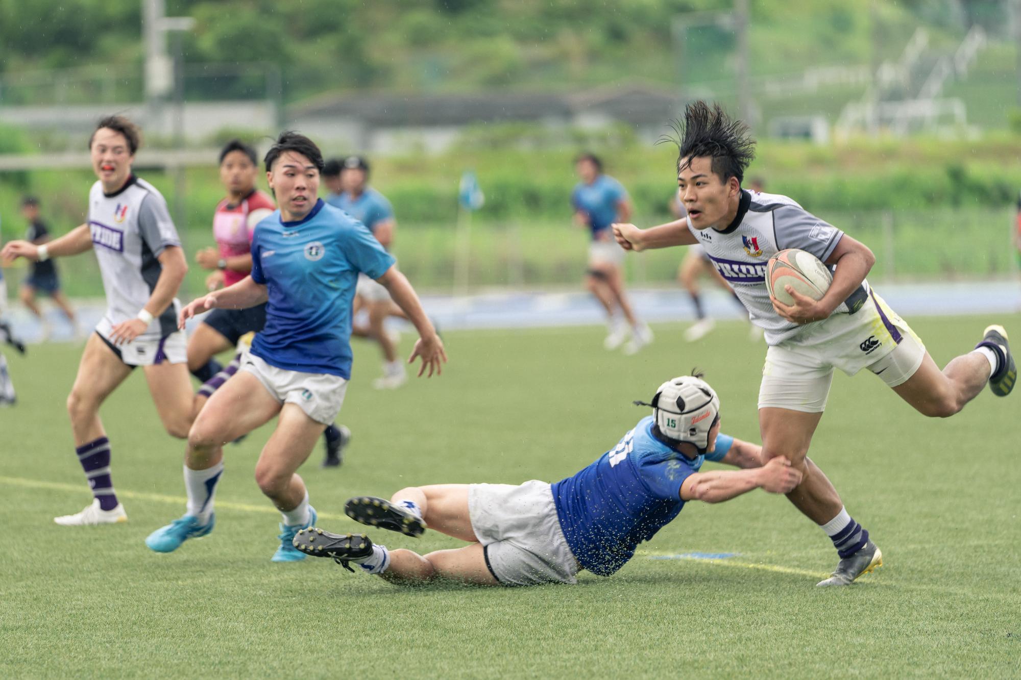 青いユニフォーム姿の選手が、ボールを持っている白いユニフォーム姿の相手選手にタックルする様子の写真