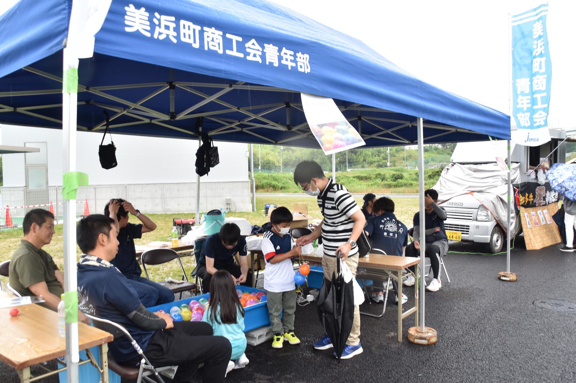 美浜町商工会青年部と書かれた青いテントブースでヨーヨー釣りを楽しむ親子の写真