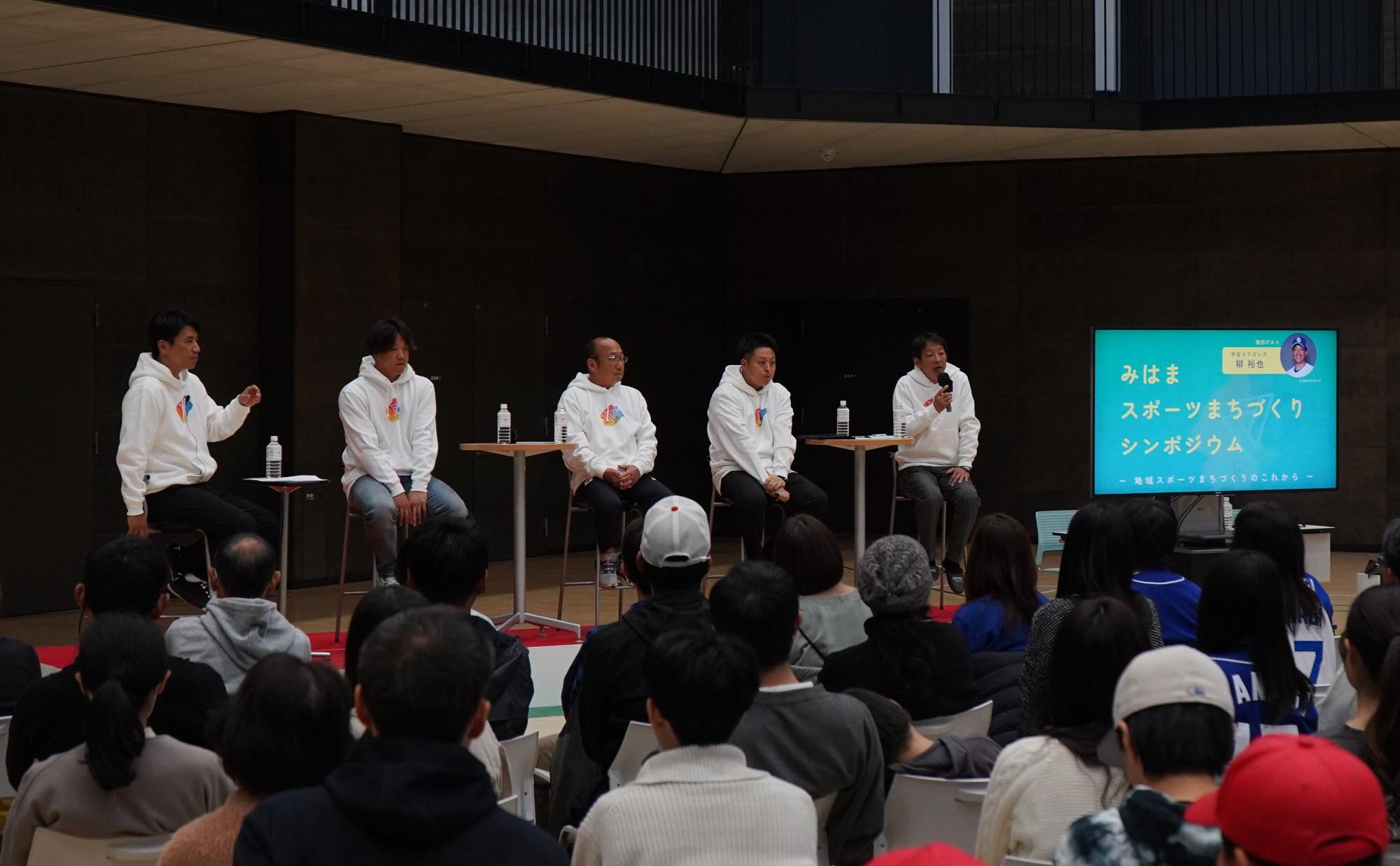 「みはまスポーツまちづくりシンポジウム」の文字がスクリーンに映し出され、椅子に座りマイクで話をする選手、その横に4名の選手が座っており、多くの参加者が聞いているシンポジウムの様子の写真
