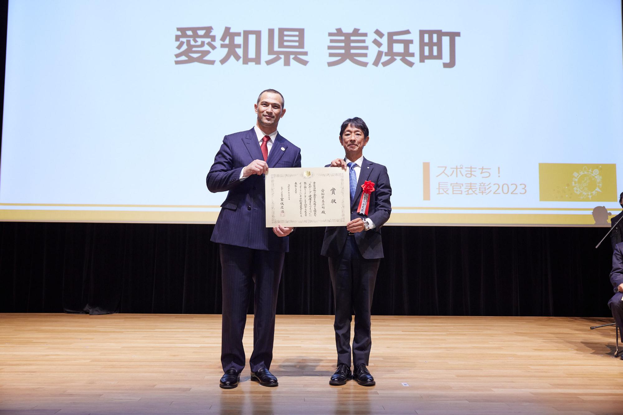スクリーンに「愛知県美浜町」が映し出され、市長とスーツを着た男性が賞状を持っている記念写真