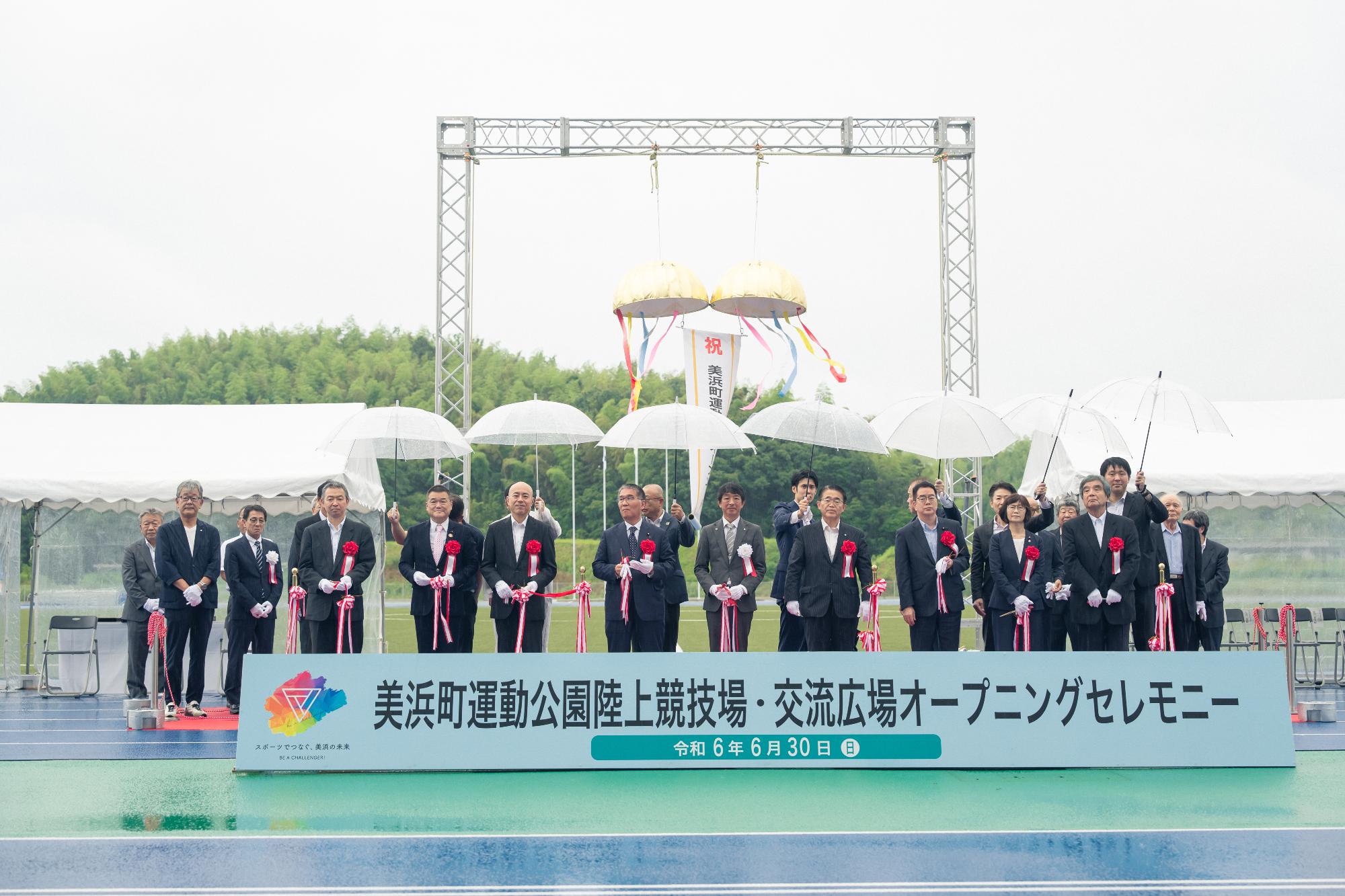地面に置かれた横長の看板に「美浜町運動公園陸上競技場・交流広場オープニングセレモニー」と書かれ、雨が降る中傘の下で愛知県知事や町長と関係者の方々がテープカットをしている写真