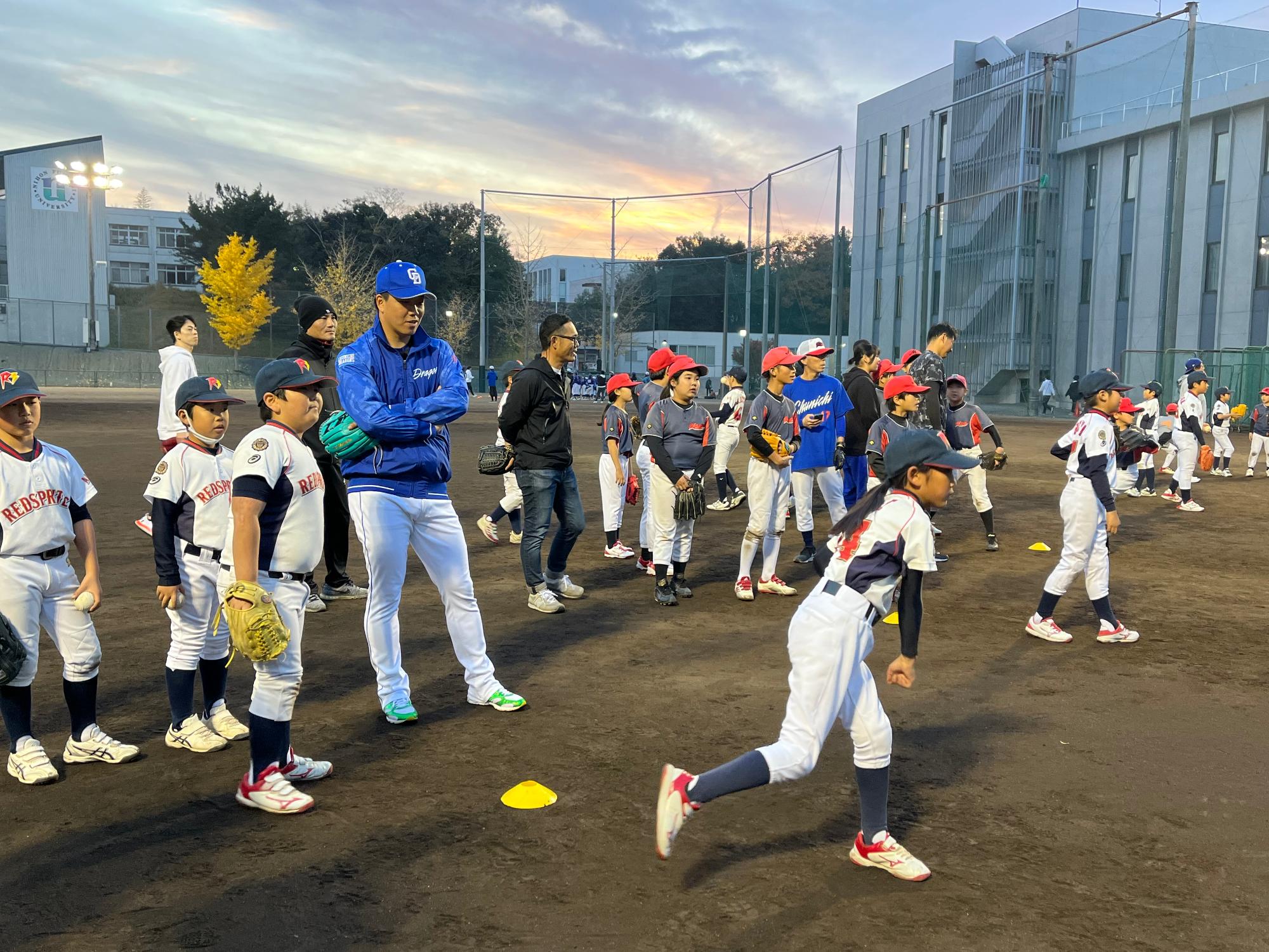 夕暮れの空のもと、柳裕也投手が見守る中、子供達がキャッチボールをしている様子の写真