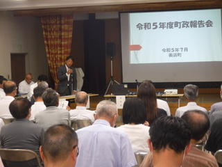 スーツを着た男性が大勢の参加者たちの前で正面のスクリーン画面に映し出された資料に沿って説明をしている写真