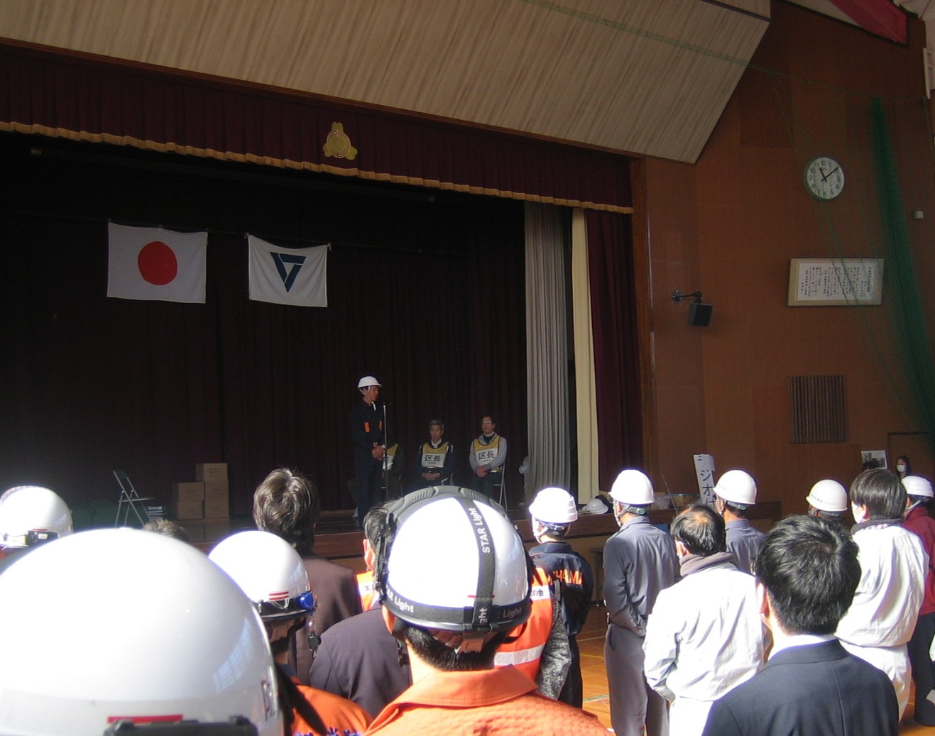 南部体育館ステージ上であいさつする八谷町長と、整列して挨拶を聞く訓練参加者。ステージ上には国旗と美浜町旗が掲げられている。